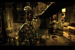 Screenshot featuring a 3 dimensional assemblage sculpture created with a small, white sculpted head and arm, red heart shaped object in the chest areal, and wire spiral below the waist. The setting is made of paper structures that resemble pages with text and with glass bulbs. The scene is bathed in a warm yellow light.