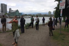 A scattered group of people stand on a beach pathway by the shore at False Creek in Vancouver. A marina with docks and highrise glass condos are on the other shore across the water.