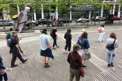 Scattered group of about 10 people walk through the stone courtyard in Olympic village at False Creek in Vancouver. A larger than life sized sparrow sculpture is in the background.
